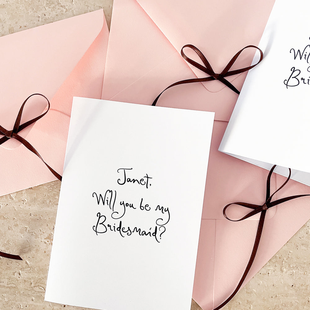 White card with 'Will you be my Bridesmaid?' text and pink envelope with a brown ribbon.