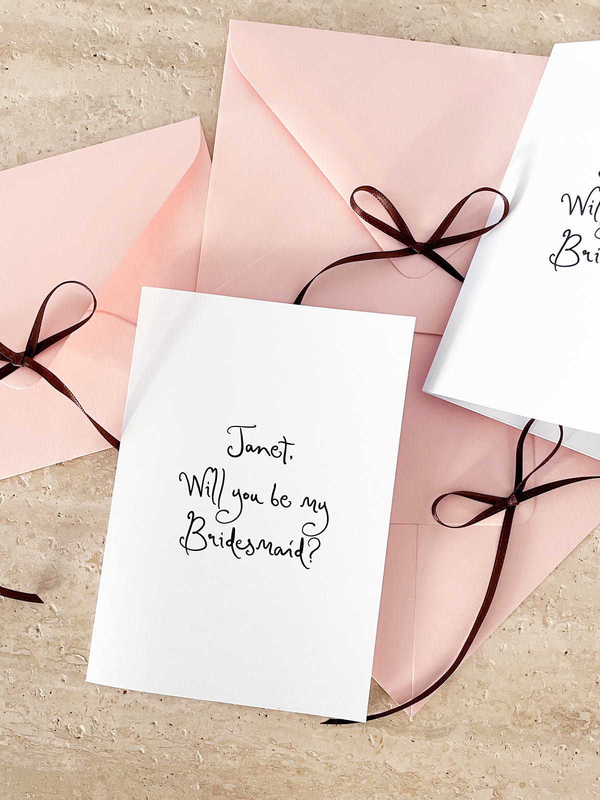 White card with 'Will you be my Bridesmaid?' text and pink envelope with a brown ribbon.